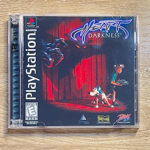 May include: A PlayStation game case for "Heart of Darkness." The cover art depicts a dark scene with a boy holding a futuristic weapon, a dog, and a shadowy figure. The game case is in a clear plastic case.