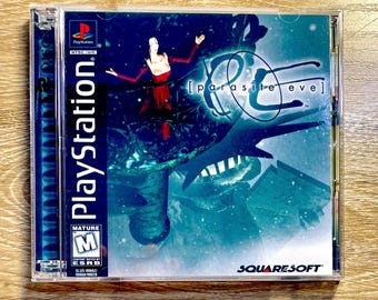 May include: A PlayStation game case for "Parasite Eve." The cover art features a woman in a red dress with outstretched arms against a blue and teal background. The PlayStation logo and "Mature" rating are visible.