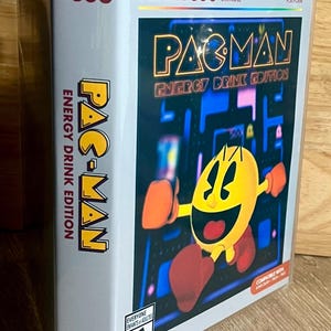 May include: An Atari 7800 video game cartridge case for Pac-Man Energy Drink Edition. The cover features a yellow Pac-Man character holding an energy drink against a dark blue maze background. The case includes the Atari 7800 logo and ESRB E rating.