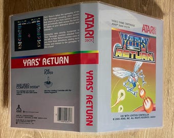 May include: A video game cartridge for the Atari 2600, titled "Yars' Return." The box features a red and white design with the game's title in bold letters. The cover art shows a futuristic insect-like creature firing a weapon. The game is for one player.