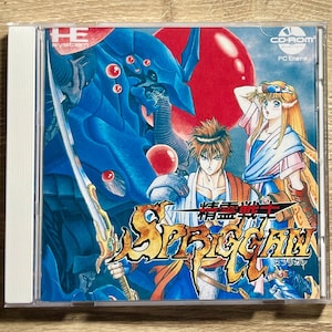 May include: A Spriggan CD-ROM game case featuring a vibrant illustration. The artwork showcases a blue monster, a warrior with a sword, and a woman. The title is in gold and orange, with Japanese text.