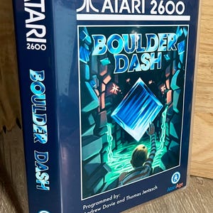 May include: A rectangular Atari 2600 Boulder Dash video game case with a blue cover featuring a character reaching for a glowing blue diamond in a cavern. The text reads Atari 2600, Boulder Dash, Programmed by: Andrew Davie and Thomas Jentzsch 2012.