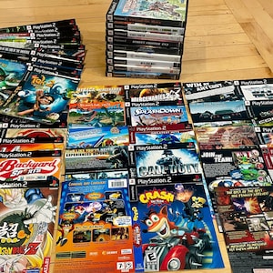 May include: A collection of PlayStation 2 video game cases and manuals arranged in a stack and spread out across a surface. The items feature various colorful graphic art, game titles, and logos, including Crash Tag Team Racing and Dragon Ball Z.