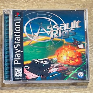 May include: A PlayStation game case for "Assault Rigs" with a futuristic vehicle design. The cover art shows a vibrant scene with a blue and green background, and the game's title is prominently displayed.