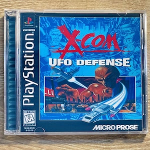 Puede incluir: Caja de juego de PlayStation para X-Com: UFO Defense. La portada presenta una "X" roja sobre el título, con imágenes de una ciudad futurista, figuras alienígenas y naves espaciales. La caja está en una funda de plástico transparente.