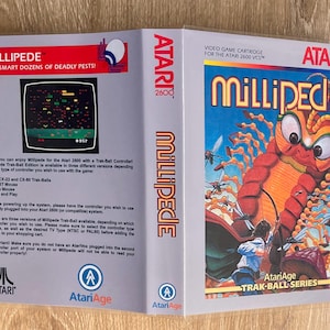 Peut inclure: Cartouche de jeu vidéo Atari 2600 Millipede. L'illustration de la boîte présente un grand mille-pattes orange sur fond de ciel bleu. Le titre du jeu est en grandes lettres stylisées. La boîte est ouverte, montrant les instructions.