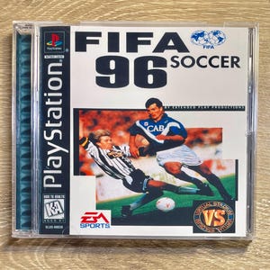 Puede incluir: Caja de juego de PlayStation para FIFA 96 Soccer. La portada presenta a dos jugadores de fútbol en acción, con el título del juego en letras blancas grandes. El logotipo de EA Sports es visible. La caja está en una funda de plástico transparente.