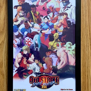 May include: A framed poster featuring a collage of characters from the video game Street Fighter III: 3rd Strike. The poster is black and white with a red, white, and blue color scheme. The text "3rd Strike Street Fighter III" is printed in the bottom center of the poster. The text "Fight for the Future" is printed in the bottom right corner of the poster.