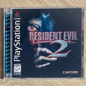 Peut inclure: Jeu PlayStation Resident Evil 2 dans un boîtier en plastique transparent. La couverture présente une image divisée d'un visage et d'une main de zombie, avec le titre du jeu en rouge et argent. Le jeu est classé M pour Mature.
