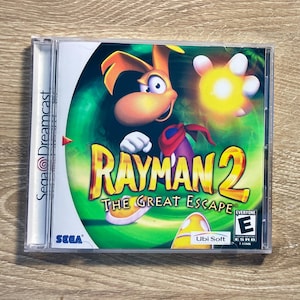 May include: A Rayman 2: The Great Escape video game case for the Sega Dreamcast. The cover art features the Rayman character with a green and yellow background. The game is rated "E" for Everyone.