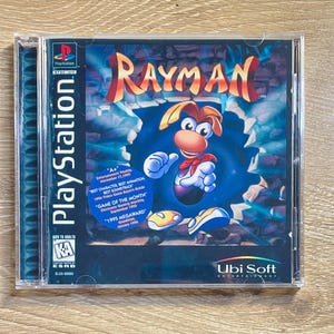 May include: A PlayStation game case for "Rayman." The cover features the game's title in orange and red, with a cartoon character in the center. The case includes text such as "Game of the Month" and "Ubi Soft Entertainment."