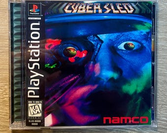 May include: A PlayStation game case for "Cyber Sled." The cover art features a futuristic, blue-toned image of a person with a robotic eye and the game's title. The PlayStation logo and the Namco logo are also visible.