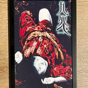 May include: Framed art print featuring a figure in a red and gold patterned garment, with a pale face and dark hair. The artwork has Japanese text and is set against a dark background. The frame is black.