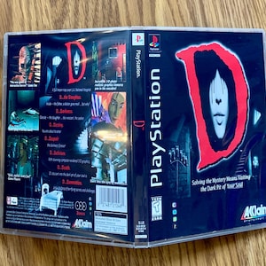 Puede incluir: Una caja de juego de PlayStation para el juego "D" con una "D" roja sobre un fondo negro. El juego está clasificado como "Teen" por la ESRB. La parte posterior de la caja presenta una descripción del juego y una lista de características. El juego es publicado por Aklaim.