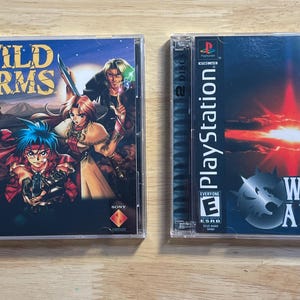May include: Two PlayStation video game cases. The first case is labeled "WILD ARMS" with a group of anime characters. The second case is labeled "WILD ARMS 2" with a wolf and anime characters. Both cases are in clear plastic.