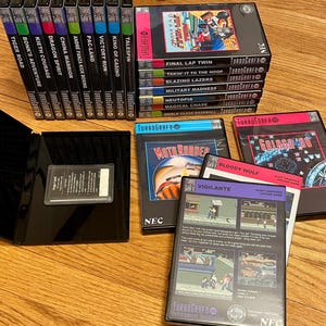 May include: A collection of TurboGrafx-16 video game cartridges, including titles like "Final Lap Twin," "Moto Roader," and "Galaga '90." The games are in their original cases, with colorful labels and graphics. The cases are black with pink and blue accents.