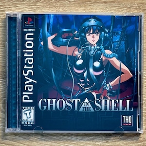 Puede incluir: Caja de juego de PlayStation para "Ghost in the Shell". La portada presenta un personaje de anime femenino en un entorno futurista con cables y tecnología. El juego está clasificado como "Teen" por la ESRB.