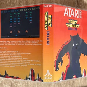 May include: Atari 2600 Space Invaders Deluxe game cartridge. The box is red with the game title and a graphic of a monster. The game's features are listed on the back of the box. The game's screen shows the classic Space Invaders game.