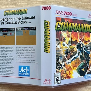 May include: A vintage Atari 7800 Commando video game cartridge box. The box art features a soldier in combat with explosions and the game title. The box is in a protective plastic case.