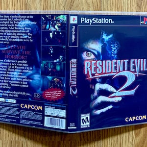 May include: A PlayStation game case for Resident Evil 2. The front of the case features a close-up of a zombie hand with a bloody mouth. The title "Resident Evil 2" is in large, white letters. The back of the case features a description of the game and a mature rating.