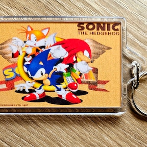 May include: A clear plastic keychain featuring a colorful illustration of Sonic the Hedgehog, Tails, and Knuckles. The image is set against a yellow background with the text "Sonic the Hedgehog" and "SEGA Enterprises Ltd. 1997".