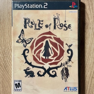 May include: A PlayStation 2 game case for "Rule of Rose." The cover art features a central rose design with a silhouette of a girl, a butterfly, a rabbit, and other decorative elements. The game is rated Mature 17+.