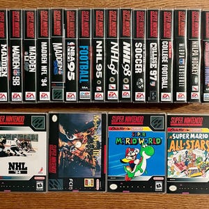 May include: A collection of Super Nintendo video game cases, including fifteen vertical spines labelled Madden, NBA, NHL, FIFA, and College Football, alongside four front-facing game boxes: NHL '94, Might and Magic III, Super Mario World, and Super Mario All-Stars.