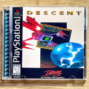 May include: A PlayStation game case for "Descent." The cover art shows a futuristic spaceship design in red and purple, emerging from a black and yellow hazard-striped portal. A blue, glowing orb is also present. The text "Interplay" and "BY GAMERS. FOR GAMERS." are visible.