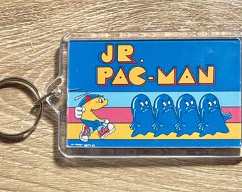 May include: A rectangular keychain featuring the text "JR. PAC-MAN" in yellow and orange on a blue background. The design includes a yellow Pac-Man character and four blue ghosts. A silver key ring is attached.