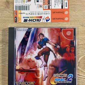 May include: A Dreamcast video game case with a red, white, and blue background. The game is called "Capcom vs. SNK 2: Millionaire Fighting 2001". The game features characters from both Capcom and SNK fighting games. The case is in good condition and has a Dreamcast logo on the front.