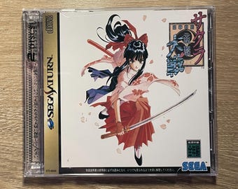 May include: A Sega Saturn video game case for Sakura Wars featuring a central illustration of a character with long black hair, wearing a pink and red outfit, and holding a katana. The cover includes Japanese text, the Sega Saturn logo, and a disc icon.