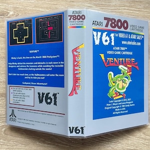 May include: Atari 7800 Venture video game cartridge case with blue and light gray panels. The front features the Venture logo, a green dragon illustration, and text including V61 Video 61 & Atari Sales. The back displays game screenshots.