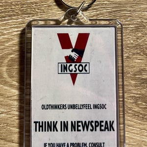 May include: A rectangular keychain with a silver ring. The keychain features a white background with the text "INGSOC" and "THINK IN NEWSPEAK". The design includes a red "V" shape with a handshake graphic.