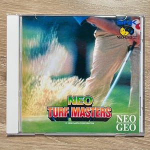 May include: A Neo Geo CD video game case for Neo Turf Masters featuring a golfer in tan trousers holding a golf club on a green course. The cover displays the title Neo Turf Masters in bold letters, the SNK logo, and the 1996 Nazca Corporation copyright.
