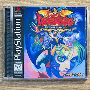 Puede incluir: Caja de juego de PlayStation para Darkstalkers: The Night Warriors. La portada presenta la cara de una mujer con ojos verdes y cabello azul, rodeada de varios personajes monstruosos en tonos azules y morados. El juego está clasificado como Teen.