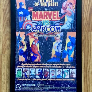 May include: A framed poster advertising the video game Marvel vs. Capcom: Clash of Super Heroes. The poster features a black background with a large, white star in the center. The words "The Best of the Best!" are at the top of the poster, and the words "Marvel vs. Capcom: Clash of Super Heroes" are in the center. The poster also features images of various Marvel and Capcom characters, including Captain America, Spider-Man, Wolverine, Ryu, and Mega Man.