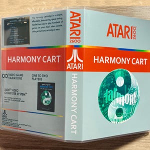 May include: A Harmony Cartridge box for the Atari 2600 video game console. The box is light blue with red and white text, including "ATARI 2600" and "HARMONY CART." A yin-yang design is featured on the front, with the Atari logo.