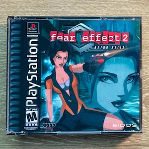 Può includere: Custodia del gioco per PlayStation di "Fear Effect 2: Retro Helix". La copertina presenta una donna in abito arancione e nero che tiene una pistola, con un volto blu sullo sfondo. Il gioco è classificato Mature.