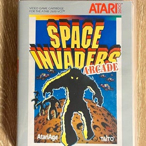 May include: A vintage Atari 2600 video game cartridge for Space Invaders Arcade. The game's cover art features a large black alien figure with yellow outlines, set against a blue sky and a brown planet surface. The game title is in yellow and red.