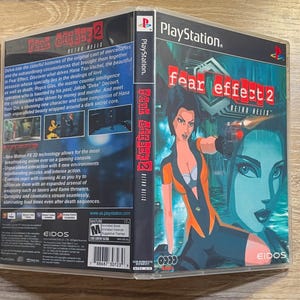 Può includere: Custodia del gioco per PlayStation di "Fear Effect 2: Retro Helix". La copertina presenta un personaggio femminile in un completo arancione e blu, con una pistola. Il titolo del gioco è in rosso e bianco. Il retro della custodia contiene dettagli e schermate del gioco.