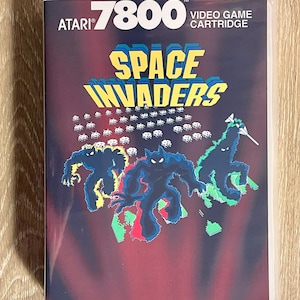 May include: Atari 7800 Space Invaders video game cartridge. The cartridge features the game's title in yellow and blue, with a background of alien figures in blue, green, and red. The Atari logo is in white.