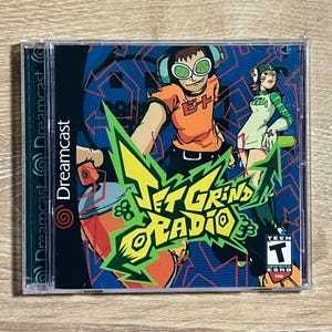 May include: A Sega Dreamcast video game case for Jet Grind Radio featuring a colorful graphic illustration of two characters with spray paint cans. The cover art displays the title in bold yellow lettering and includes a Teen ESRB rating icon.