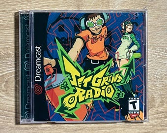 May include: A Sega Dreamcast video game case for Jet Grind Radio featuring a colorful graphic illustration of two characters with spray paint cans. The cover art displays the title in bold yellow lettering and includes a Teen ESRB rating icon.