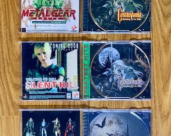 May include: Four PlayStation game cases with the titles "Metal Gear Solid", "Castlevania Symphony of the Night", "Silent Hill", and "Castlevania Symphony of the Night". The cases are on a wooden surface.