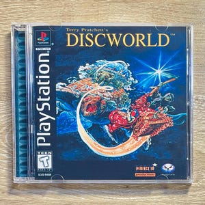 Puede incluir: Caja de juego de PlayStation para "Discworld" de Terry Pratchett. La portada presenta una escena fantástica con un personaje montando un vehículo, sobre un fondo azul y cósmico. La caja es de plástico transparente.