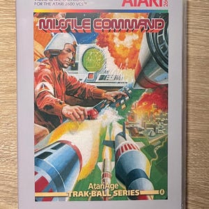 May include: A rectangular video game cartridge case for the Atari 2600 VCS titled Missile Command. The cover art features a person in a red uniform operating a control console with a trackball, surrounded by missiles and explosions in a city.