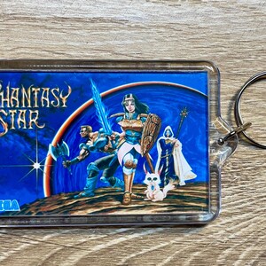 May include: A rectangular keychain featuring the "Phantasy Star" video game cover art. The artwork depicts a female warrior with a sword, a male warrior with an axe, and a robed figure, all set against a blue space background. The SEGA logo is visible.
