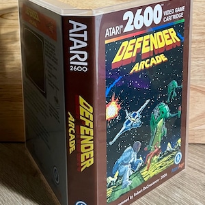 May include: A clear plastic video game case for the Atari 2600 game Defender Arcade. The cover features a space battle illustration with a spaceship, a green alien craft, and a figure on a planet surface. The spine displays the Atari 2600 logo.