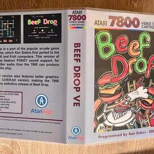 May include: Atari 7800 video game cartridge for Beef Drop. The game's cover art features a cartoon burger and a chef character. The game is programmed by Ken Siders in 2006. The cartridge is in a clear plastic case.