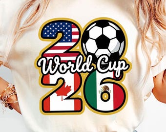 Op de afbeelding: Crèmekleurig T-shirt met een voetbal en de tekst "World Cup 26". De cijfers "2" en "6" zijn ontworpen met de vlaggen van de Verenigde Staten, Canada en Mexico. Het shirt heeft een casual, sportieve stijl.